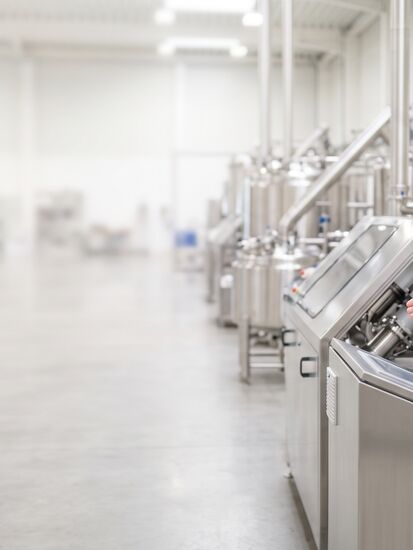 Zwei Mitarbeitende in Laborkitteln mit Schutzbrille stehen an einem Bedienpult in einer Produktionshalle mit Edelstahlrohren