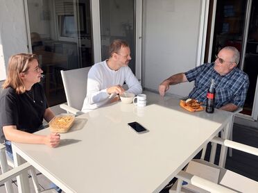 Drei Personen sitzen auf der Terrasse von agorum beim Mittagessen und sprechen, auf dem Tisch liegt das Smartphone, das die Aufnahme mitschreibt