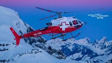 Ein Hubschrauber der Swiss Helicopter Group fliegt über schneebedeckte Berge bei Sonnenuntergang.