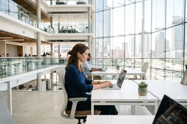 Mitarbeiterin arbeitet konzentriert am Laptop in einem hellen Großraumbüro mit Glasfront und Blick auf die Stadt