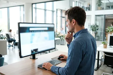 Mitarbeiter sucht nachdenklich an einem Monitor im hellen Büro, das Suchfeld oben auf dem Bildschirm im Blick