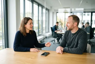 Zwei Personen im Gespräch an einem hellen Besprechungstisch, auf dem Tisch liegt ein Smartphone, niemand schreibt mit