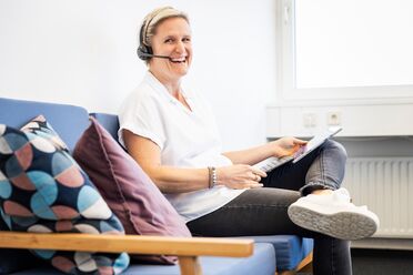 Eine Mitarbeiterin sitzt auf einem blauen Sofa im Büro und arbeitet mit einem Tablet, während sie ein Headset trägt.