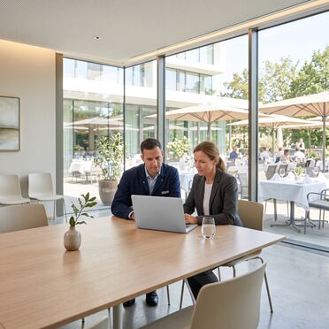 Zwei Personen am Laptop in einem Hotel, dahinter die Terrasse mit eingedeckten Tischen