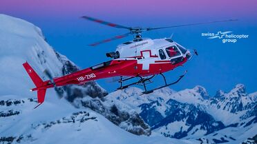 Ein Hubschrauber der Swiss Helicopter Group fliegt über schneebedeckte Berge bei Sonnenuntergang.